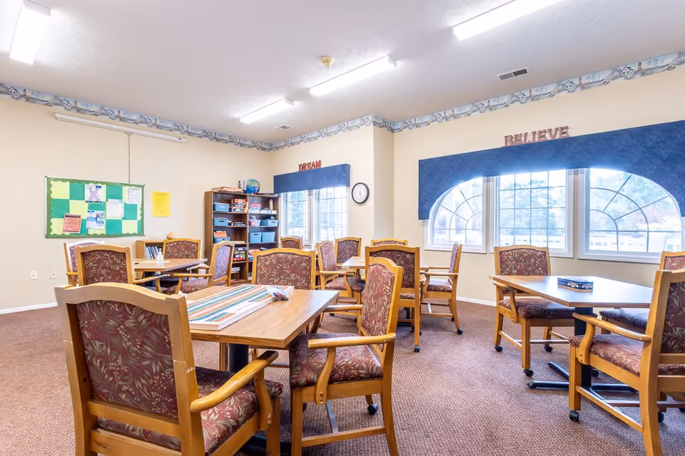 A bright room with multiple wooden tables and cushioned chairs arranged for group seating. The walls are light-colored with a decorative border near the ceiling. There are two large windows with blue valances, one with the word 'BELIEVE' above it and the other with the word 'DREAM'. A bulletin board with various papers is on one wall, and a wooden shelving unit with games and supplies is in the corner. The room has a carpeted floor and fluorescent ceiling lights.