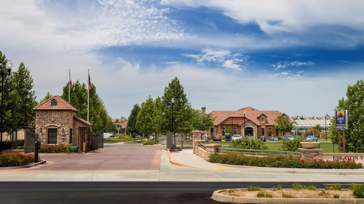 Entrance to Eskaton Village Roseville featuring a stone guardhouse, gated driveway, landscaped greenery, and a building with a red-tiled roof in the background under a partly cloudy sky.