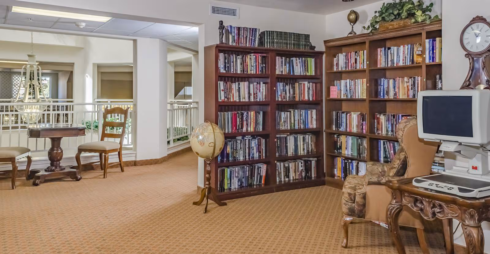 Carpeted communal library area with tall bookshelves, upholstered chairs, a globe, a small table with chairs, and a computer workstation.