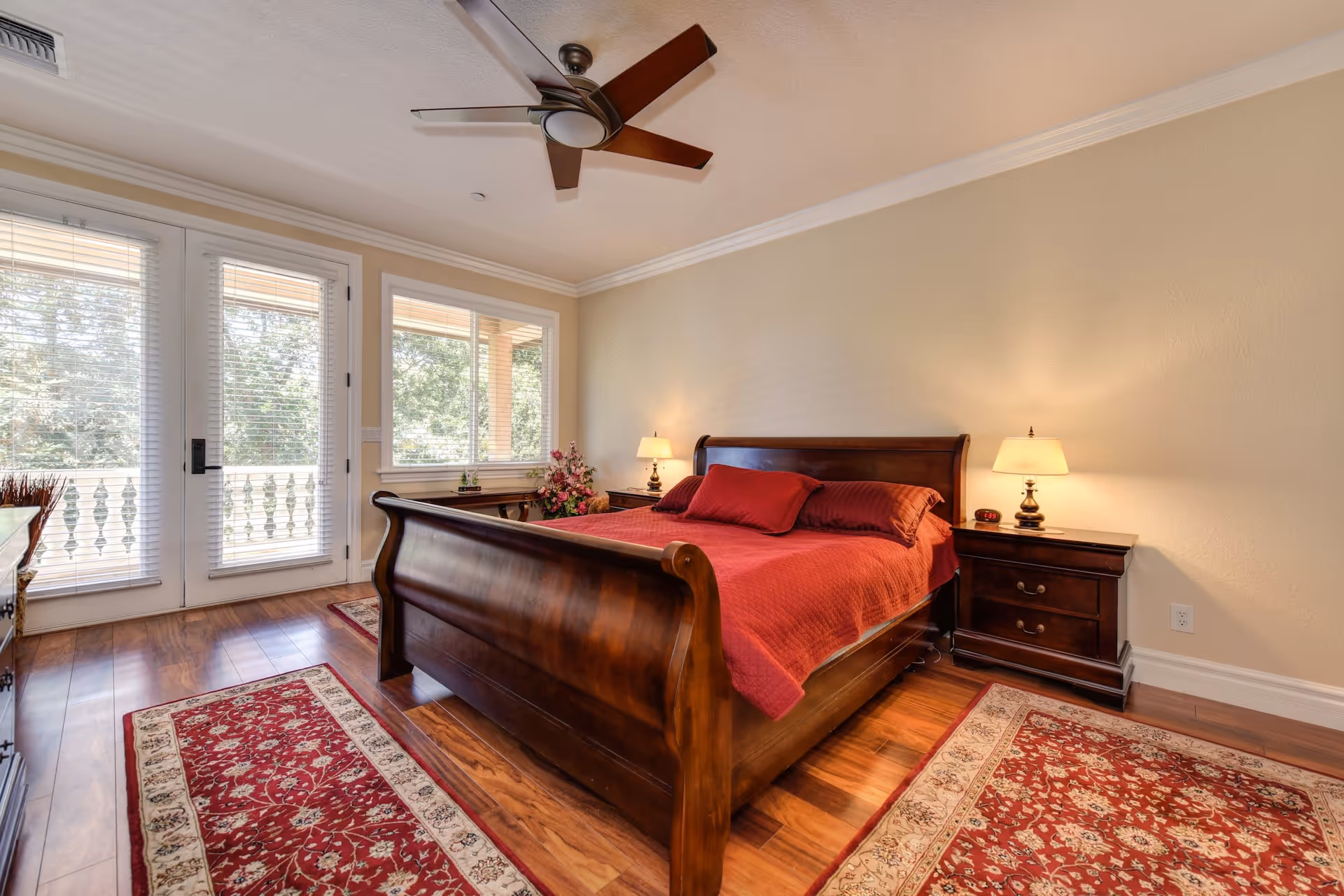 A well-lit bedroom with a wooden sleigh bed covered in a red bedspread and matching pillows. The room features wooden flooring with two red patterned rugs, a ceiling fan, two bedside tables with lamps, and large windows and glass doors leading to a balcony with white railings and greenery outside.
