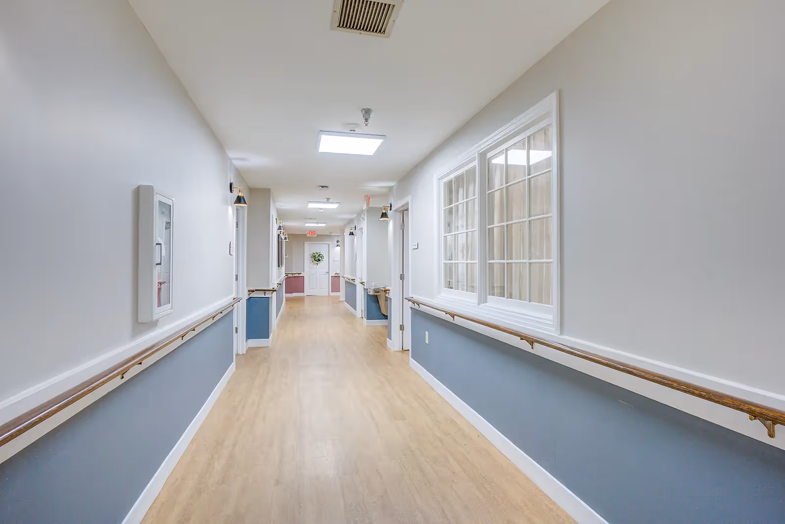 A long, well-lit hallway in a senior living facility with light wood flooring, white and blue walls, handrails on both sides, several doors along the corridor, and a window on the right side. The hallway has ceiling lights and an exit door with a wreath at the far end.