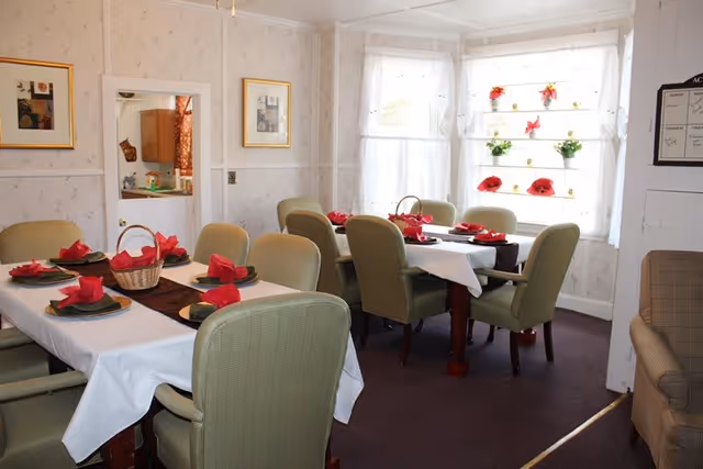 A dining room with two tables set for a meal, each covered with white tablecloths and brown runners. Each place setting has a plate with a red folded napkin. The room has light-colored wallpaper, framed pictures on the walls, and a window with sheer white curtains decorated with small flower pots. There are green upholstered chairs around the tables and a beige armchair near the doorway.