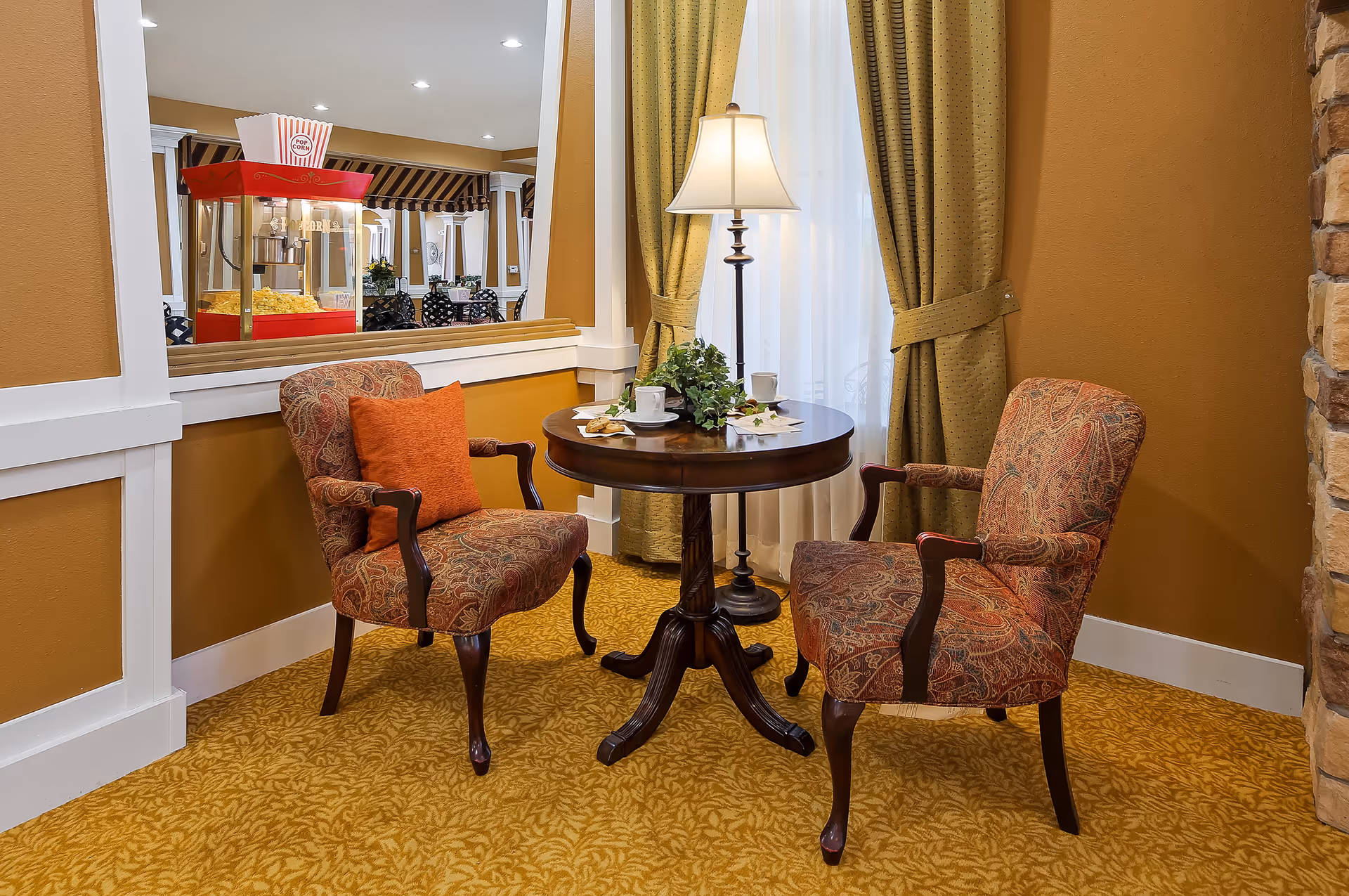 A cozy seating area with two upholstered armchairs featuring a paisley pattern and an orange cushion on one chair. Between them is a round wooden table with a small plant, two cups and saucers, and some napkins. Behind the table is a floor lamp with a white shade, and the window is dressed with green curtains. A popcorn machine and additional seating are visible through a large interior window. The room has warm yellow walls and patterned carpet.