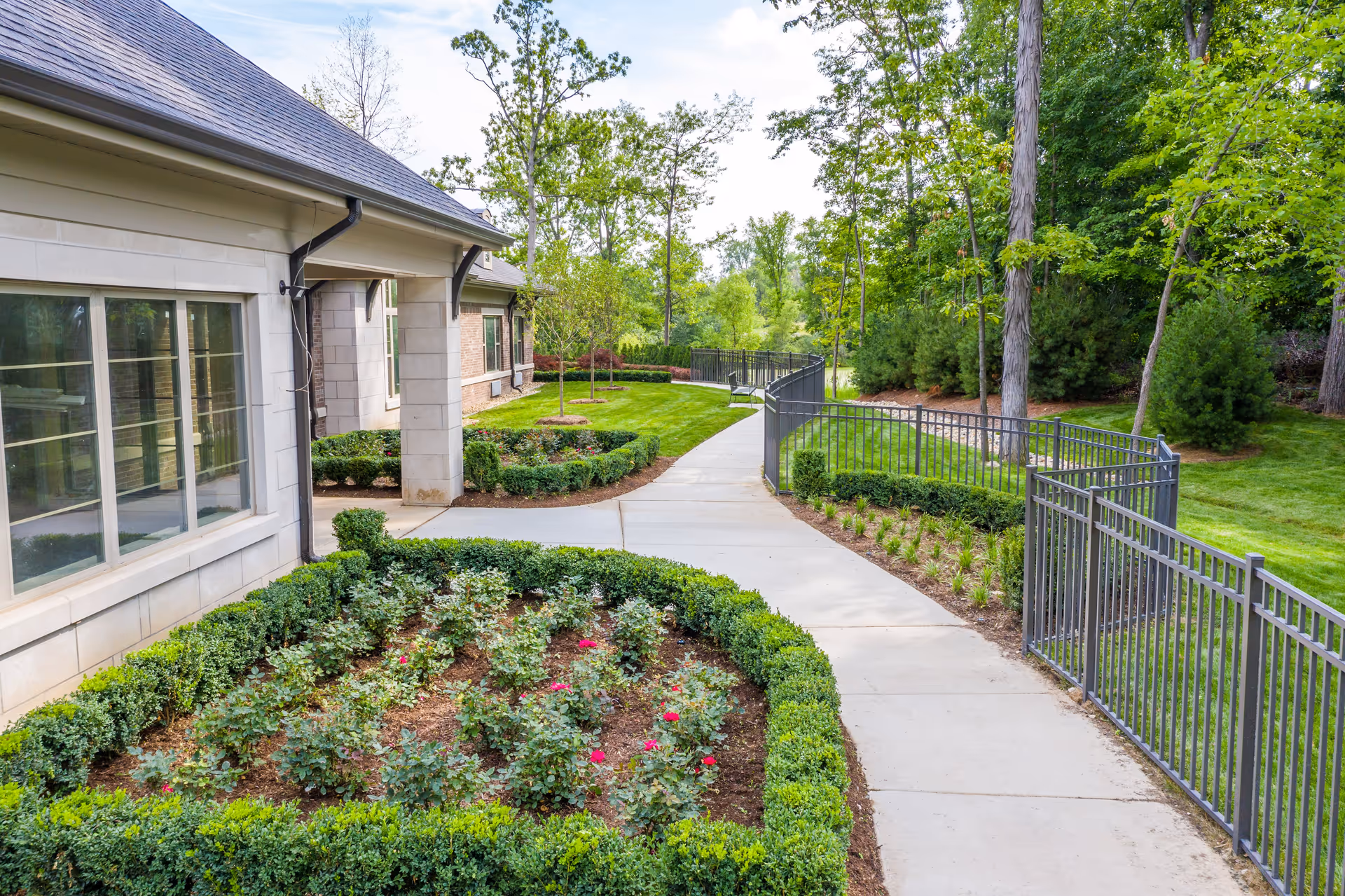 A paved walkway curves past landscaped flower beds beside a light-colored building, bordered by a metal fence and trees.