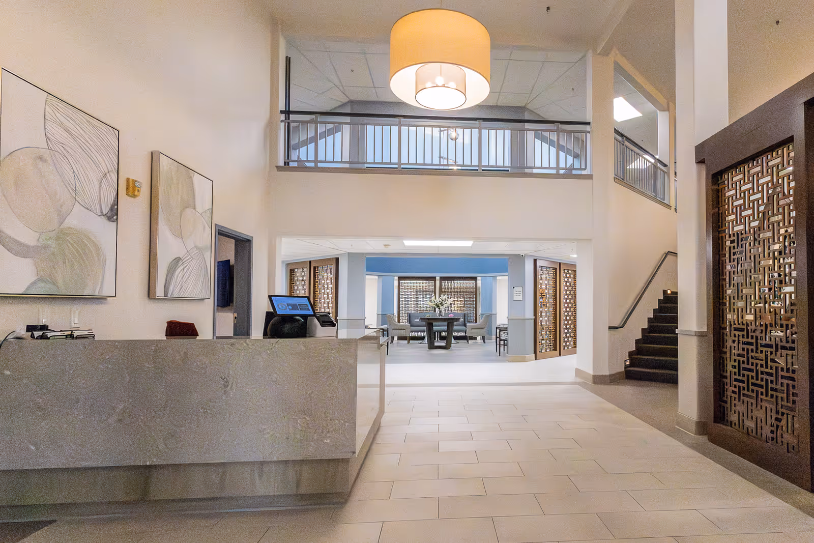 Interior view of a senior living facility lobby with a modern reception desk on the left, abstract artwork on the wall, a large hanging light fixture, and a seating area with chairs and a table in the background. There is a staircase on the right and decorative wooden panels near the entrance to the seating area.