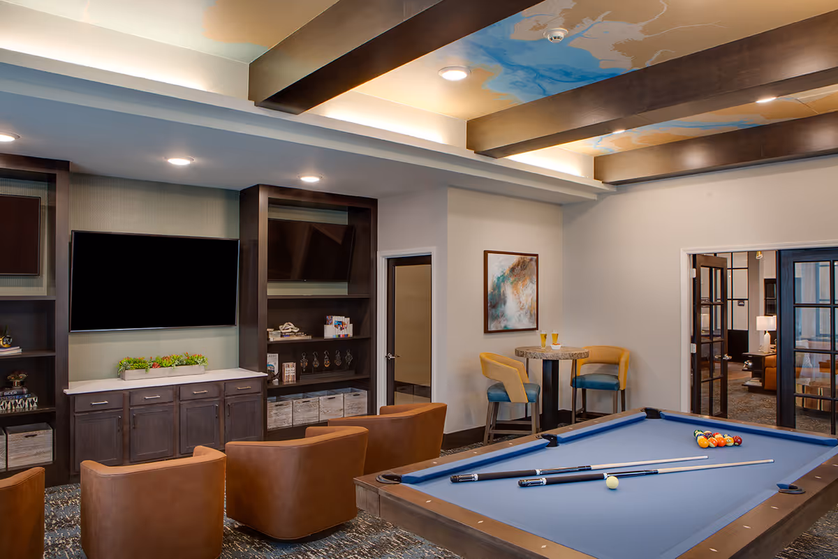 A cozy recreational room with a blue pool table in the foreground, two brown leather chairs facing a wall-mounted flat screen TV, and a small round table with two yellow and blue chairs. The ceiling has wooden beams with a decorative painted design, and there are shelves and cabinets along the wall.