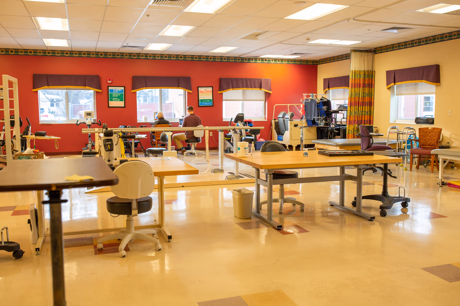 A spacious rehabilitation or physical therapy room with exercise equipment, parallel bars, tables, chairs, and two people seated near the windows working or exercising. The room has a red and beige color scheme with large windows letting in natural light.