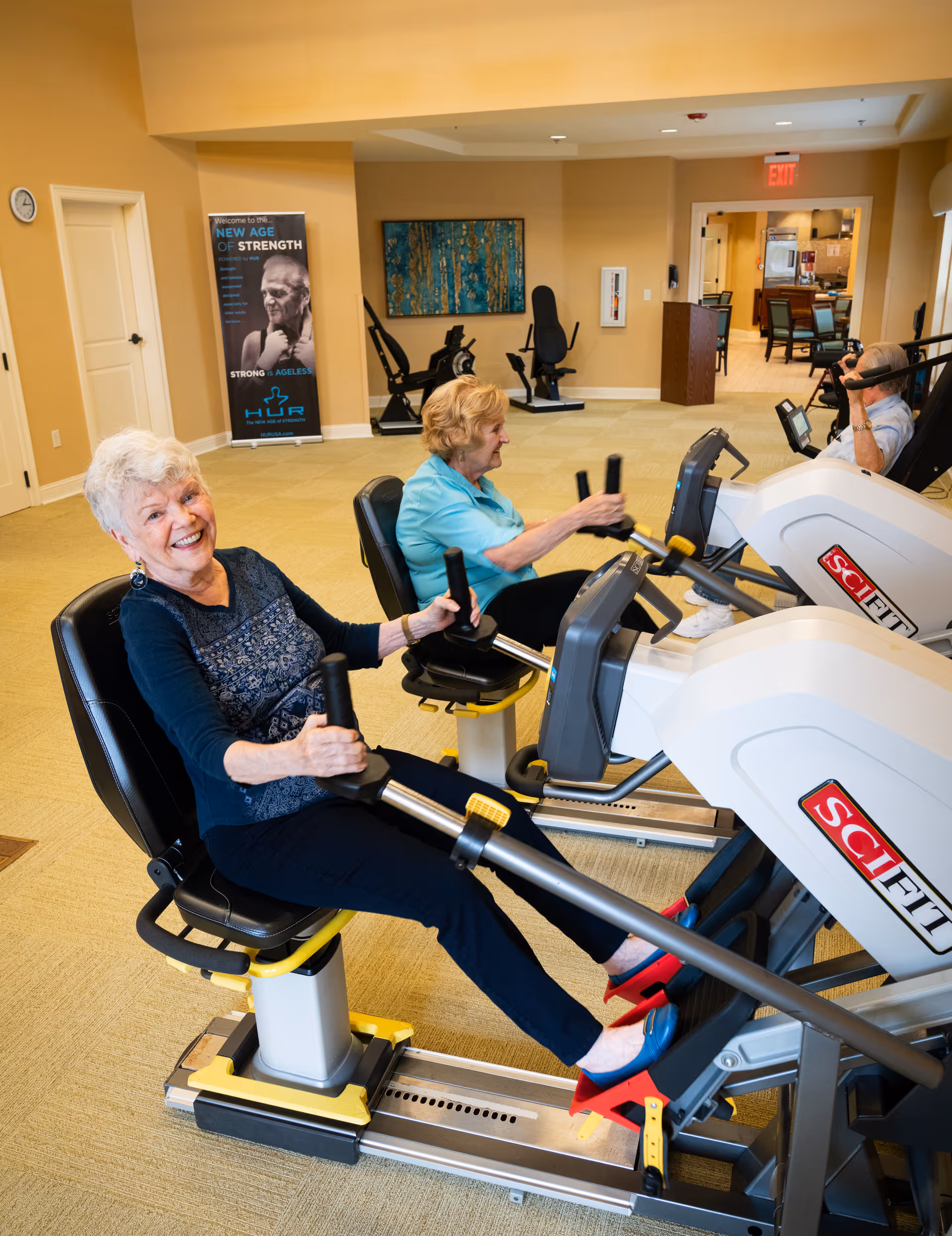 Three elderly individuals exercising on seated rowing machines in a fitness room with beige walls and carpet. One woman in the foreground is smiling at the camera, while the other two focus on their workout. There is a poster on the wall and exercise equipment in the background.