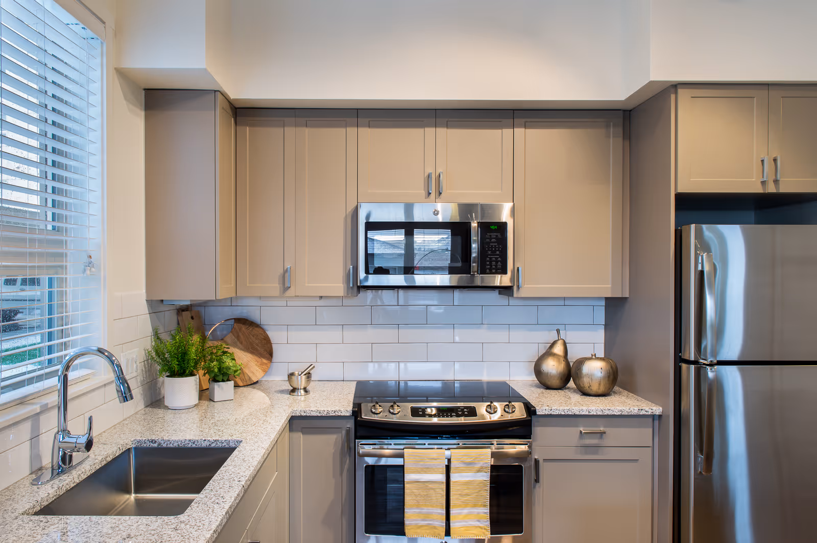 Modern kitchen with beige cabinets, a stainless steel microwave above a stove with two yellow-striped towels hanging on the oven door, a stainless steel refrigerator, a sink under a window with blinds, and decorative items including two metallic pears and potted plants on the countertop.