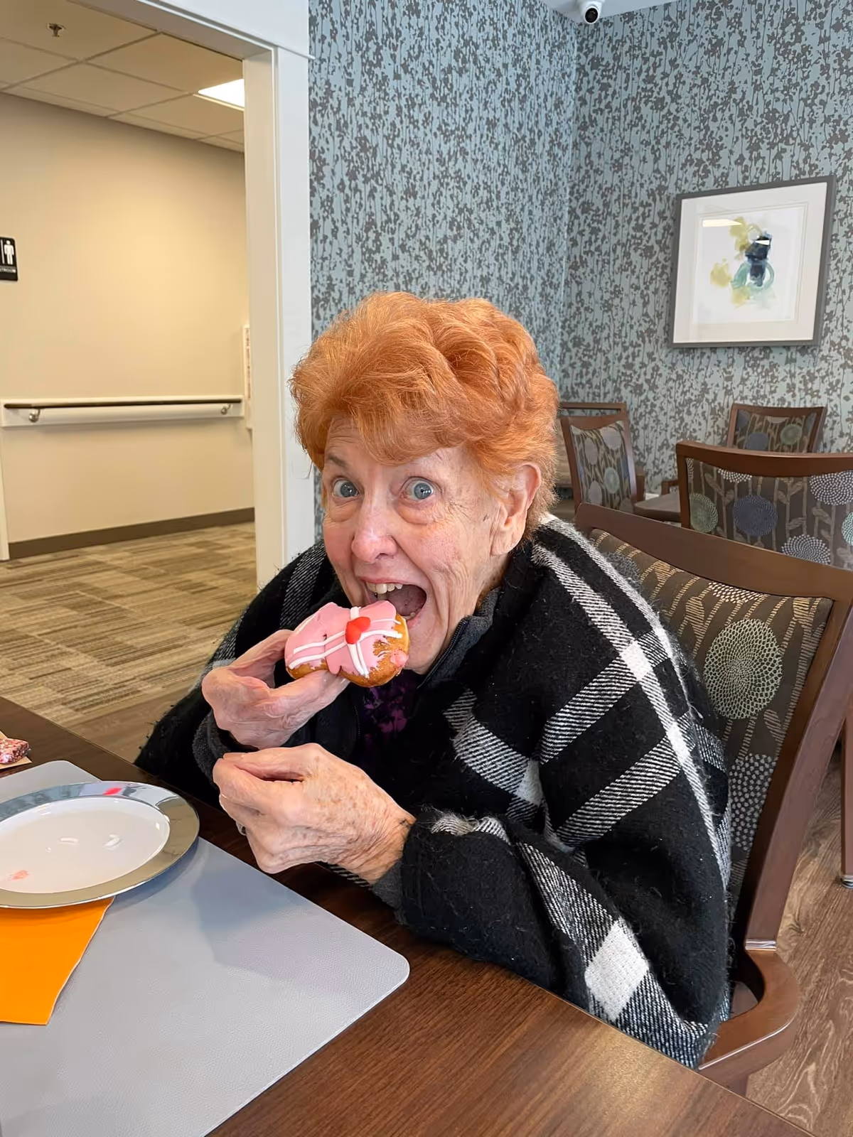 An elderly woman with red hair wrapped in a black and white plaid shawl is sitting at a table in a dining area, about to eat a pink frosted donut with a red heart decoration. The background shows patterned wallpaper, a framed abstract painting, and several chairs.