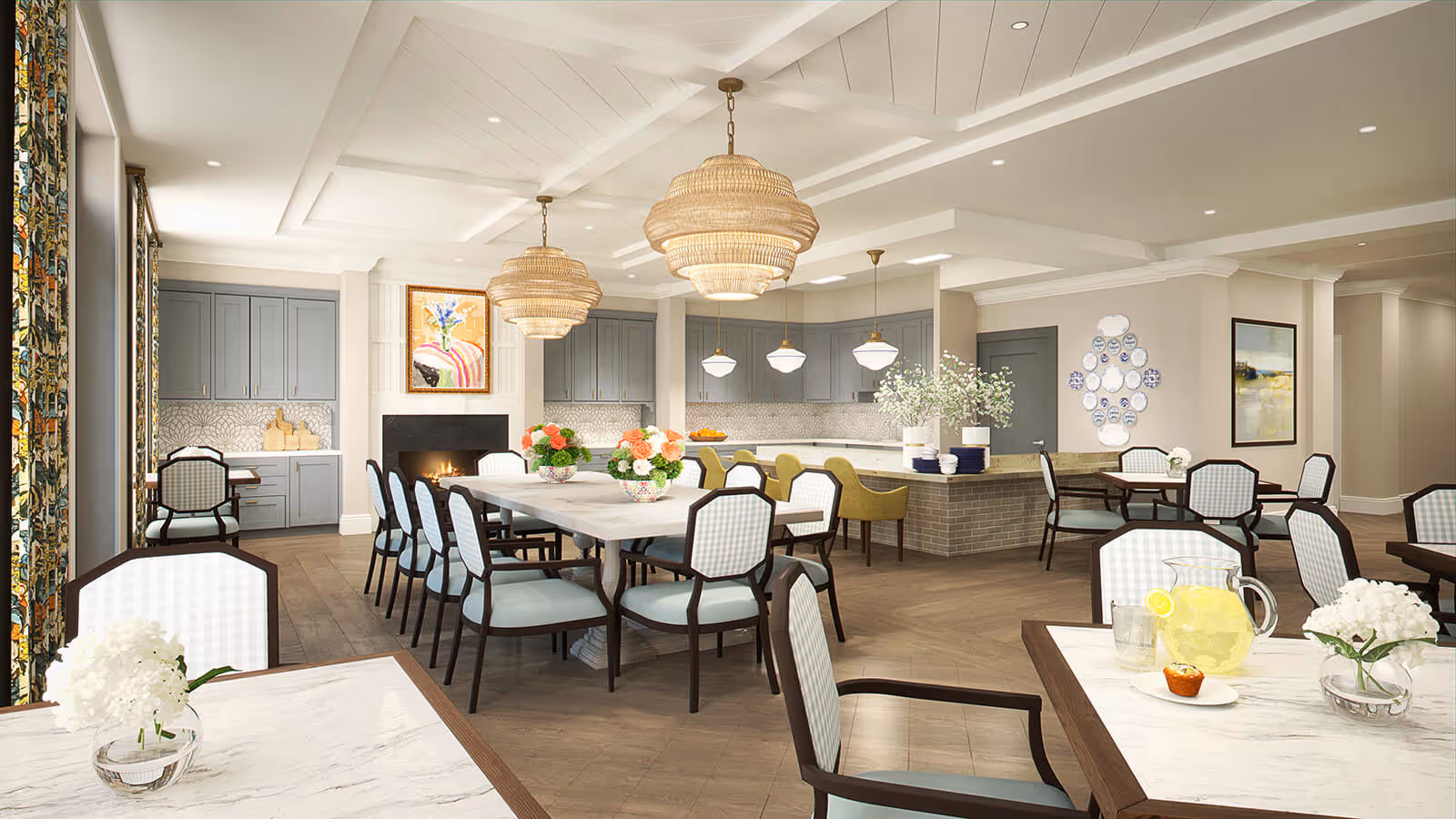 Bright communal dining room with long tables and chairs, a kitchen island with pendant lights, decorative chandeliers, and floral centerpieces.