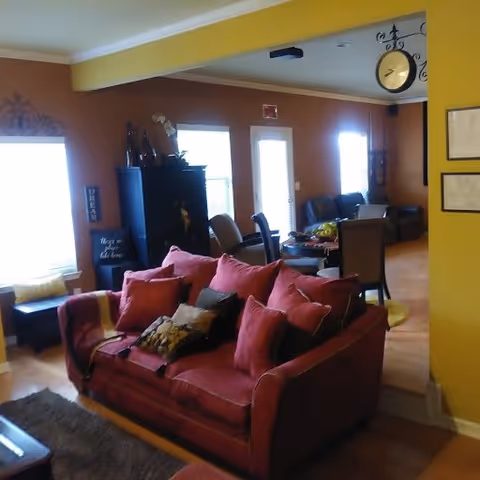 Interior view of a living room area with a red sofa adorned with multiple cushions in the foreground. Behind the sofa, there is a dining table with chairs and a dark cabinet against the wall. The walls are painted in warm tones of yellow and brown, with windows allowing natural light to enter. A decorative clock hangs from the ceiling near the transition between rooms.