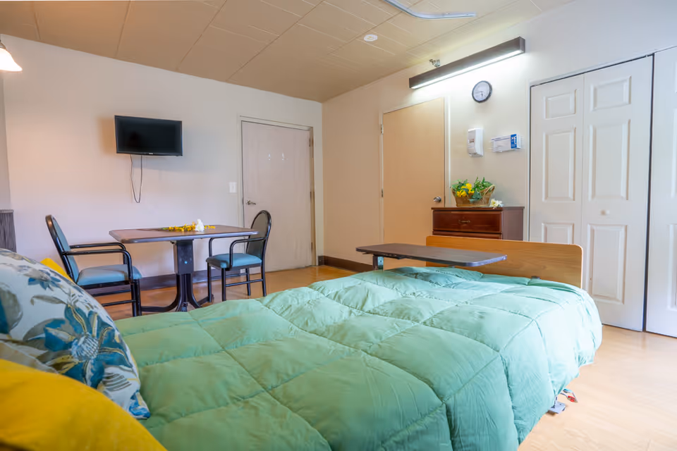 A nursing facility room with a bed covered in a green comforter and floral pillows. There is a small table with two chairs against the wall, a wall-mounted TV above the table, a wooden dresser with a basket of flowers, a clock on the wall, and two closed doors including a closet with white folding doors.