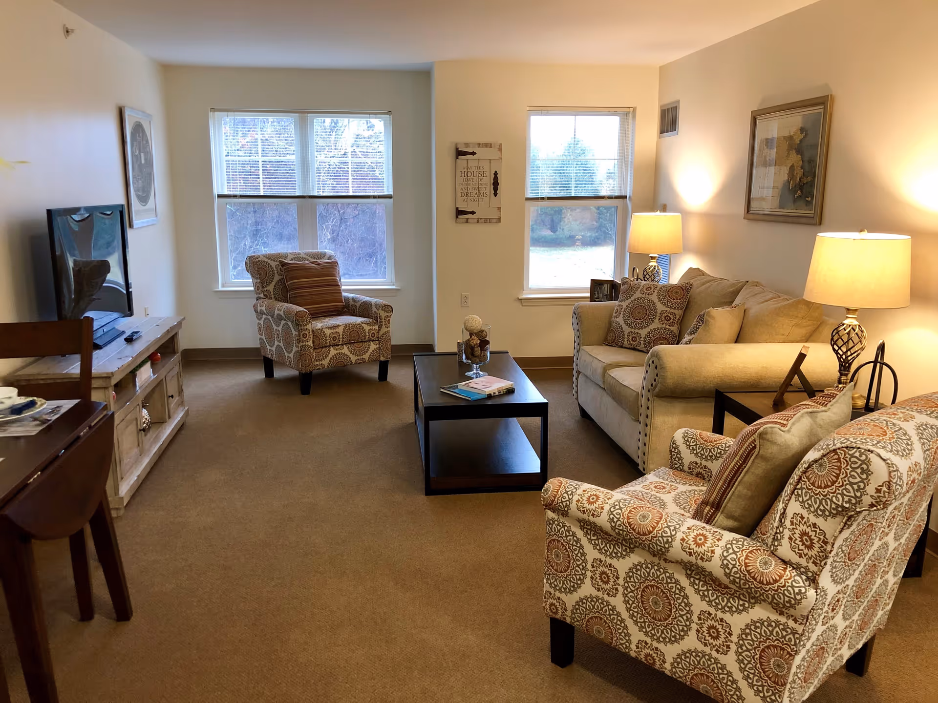 A cozy living room with beige walls and carpet, featuring a beige sofa with patterned cushions, two matching patterned armchairs, a dark wood coffee table with books and decorative items, a wooden TV stand with a flat-screen TV, two table lamps, and two windows letting in natural light.