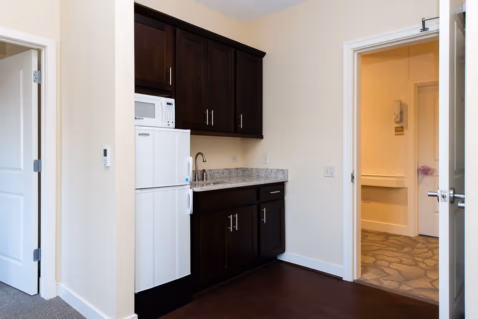 Small kitchenette area with dark wood cabinets, a white refrigerator, a microwave, and a sink with a granite countertop. Two open doors lead to adjacent rooms with beige walls and carpeted floors.