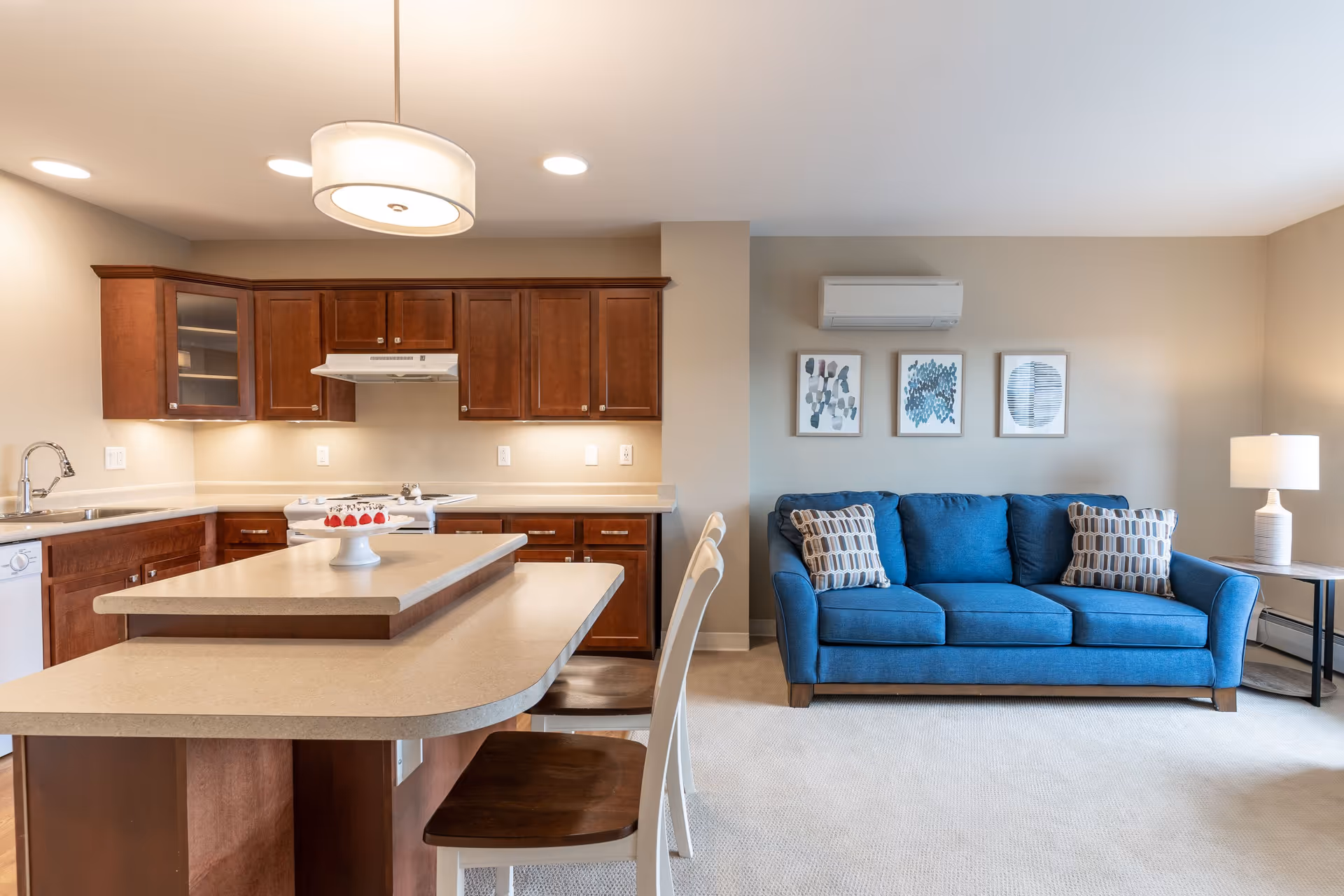 A modern open-concept living space featuring a kitchen and living room. The kitchen has wooden cabinets, a stove with a range hood, a dishwasher, and a large island with two chairs. The living room area includes a blue sofa with patterned pillows, three framed abstract artworks on the wall, a side table with a lamp, and a wall-mounted air conditioning unit.