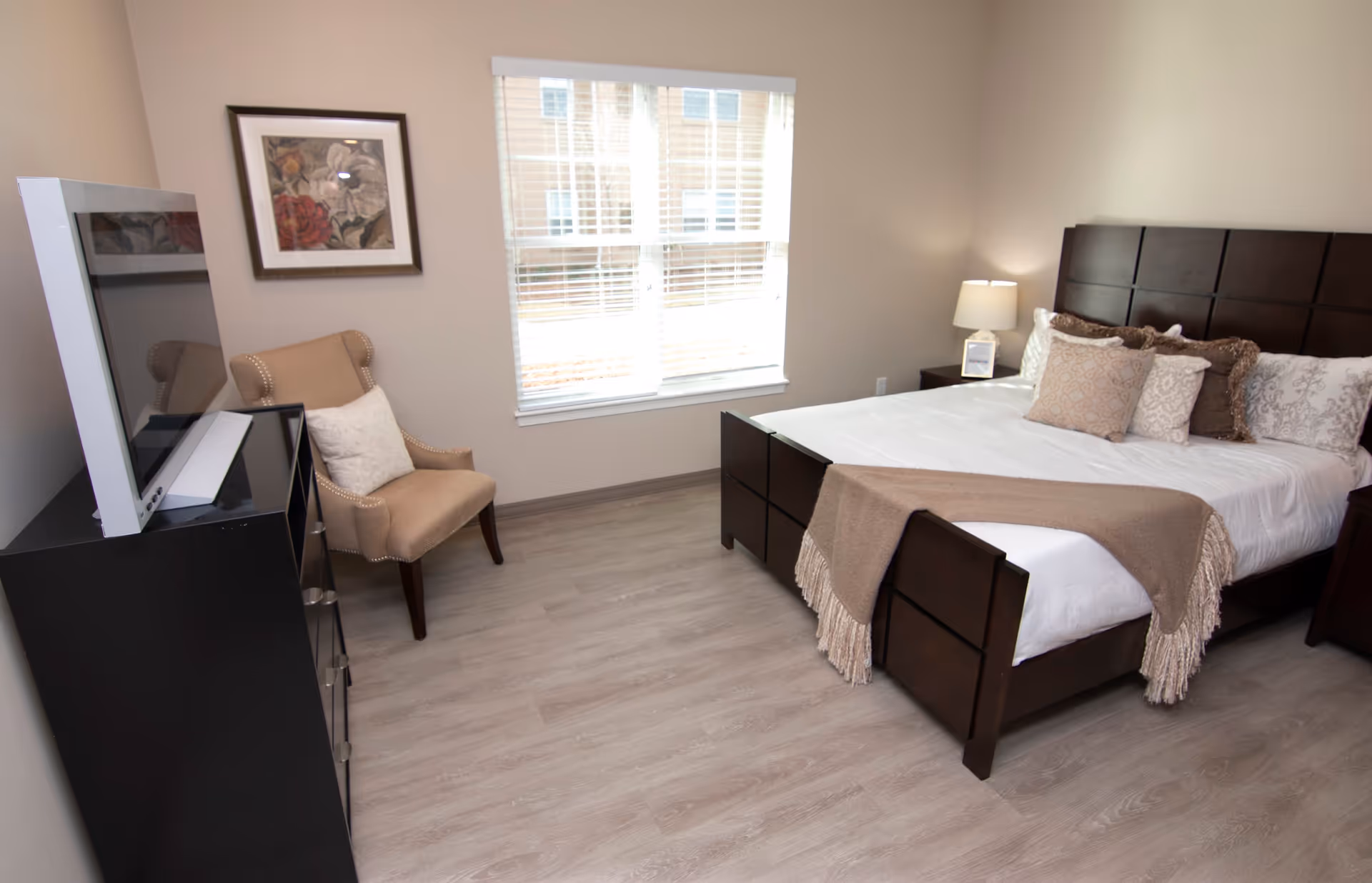 Well-lit furnished bedroom with a bed, headboard and throw pillows, bedside lamp, chair, dresser with a TV, and a window.