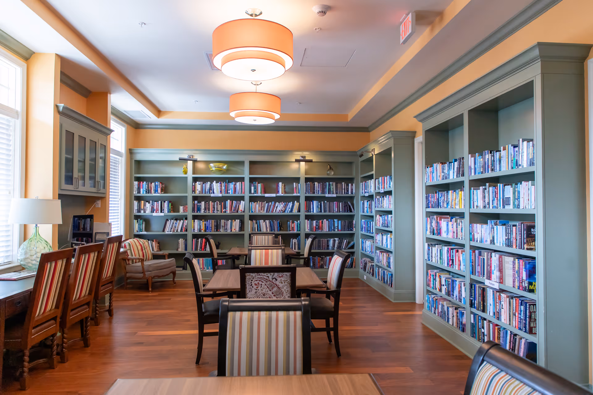 A bright and cozy library room with large windows on the left, green bookshelves filled with books along the walls, wooden tables and chairs with striped upholstery, and two large round ceiling lights.