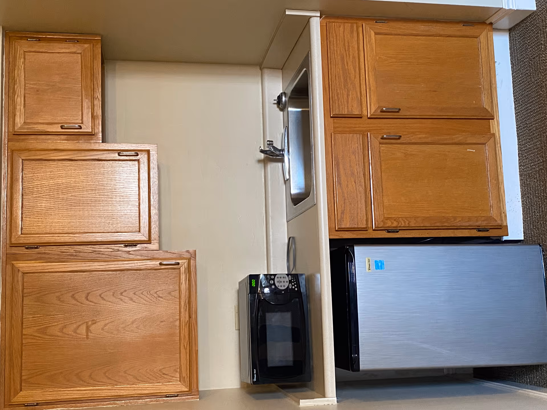 A small kitchenette area with wooden cabinets above and below a beige countertop. On the countertop, there is a black microwave oven and a stainless steel sink with a faucet. Below the countertop, there is a compact stainless steel refrigerator. The walls are painted beige and the floor is carpeted.