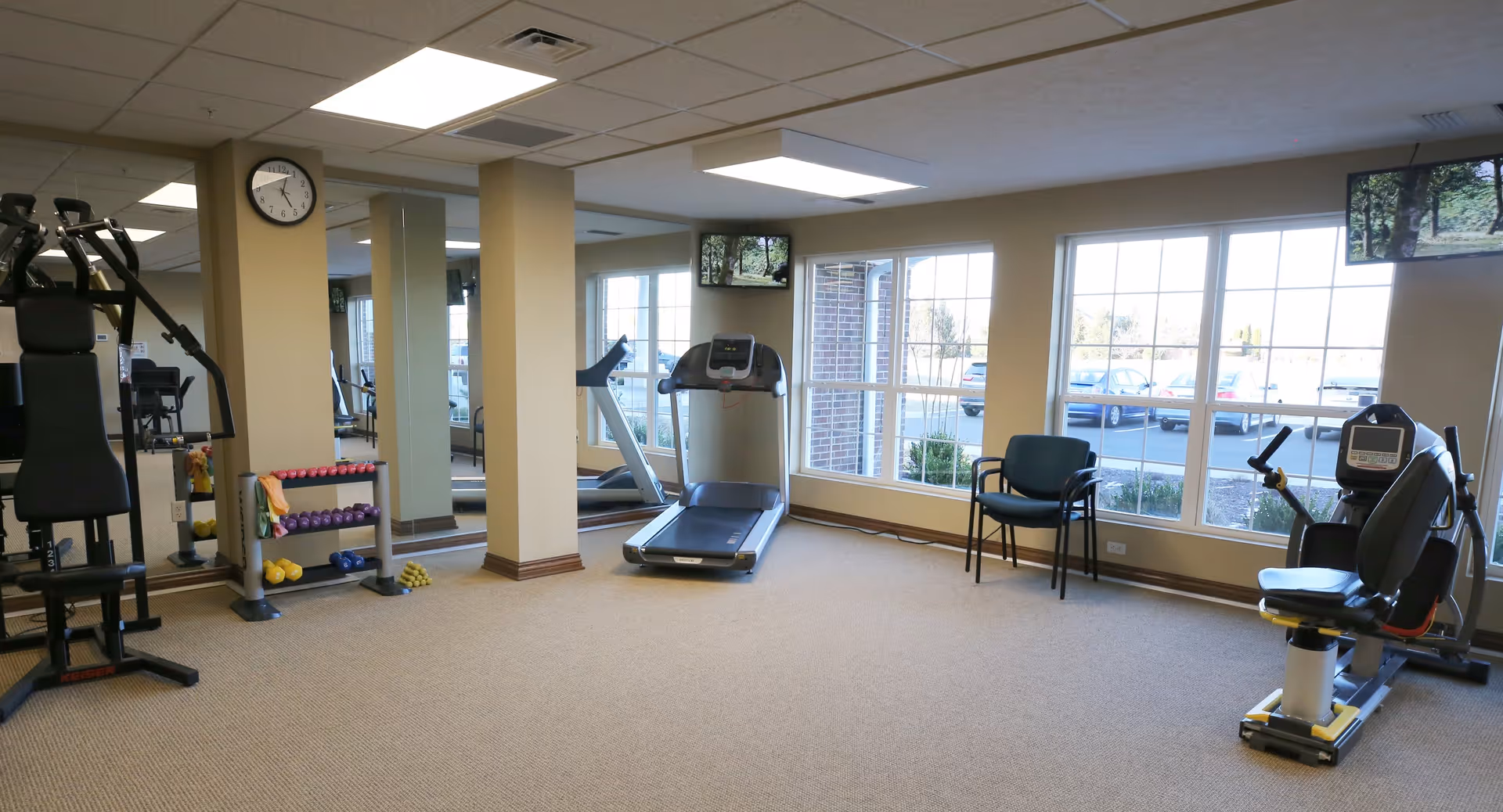 A well-lit exercise room with large windows showing a parking lot outside. The room contains various exercise equipment including a treadmill, weight machine, stationary bike, and a rack of colorful dumbbells. There are two chairs near the windows and a wall clock above a mirrored wall.