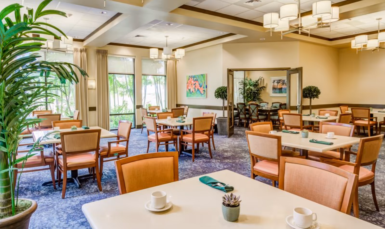 Bright dining room with multiple tables and chairs, potted plants, large windows, and pendant lighting.