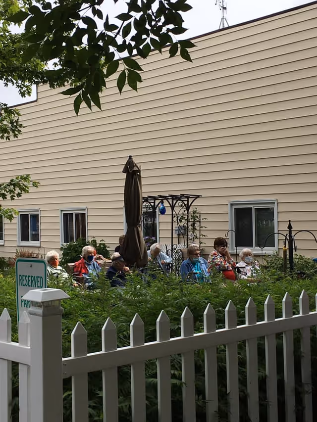A group of elderly people sitting outdoors in a garden area next to a beige building with several windows. They are seated behind a white picket fence with green bushes in front. There is a reserved parking sign and a closed patio umbrella visible among the group.