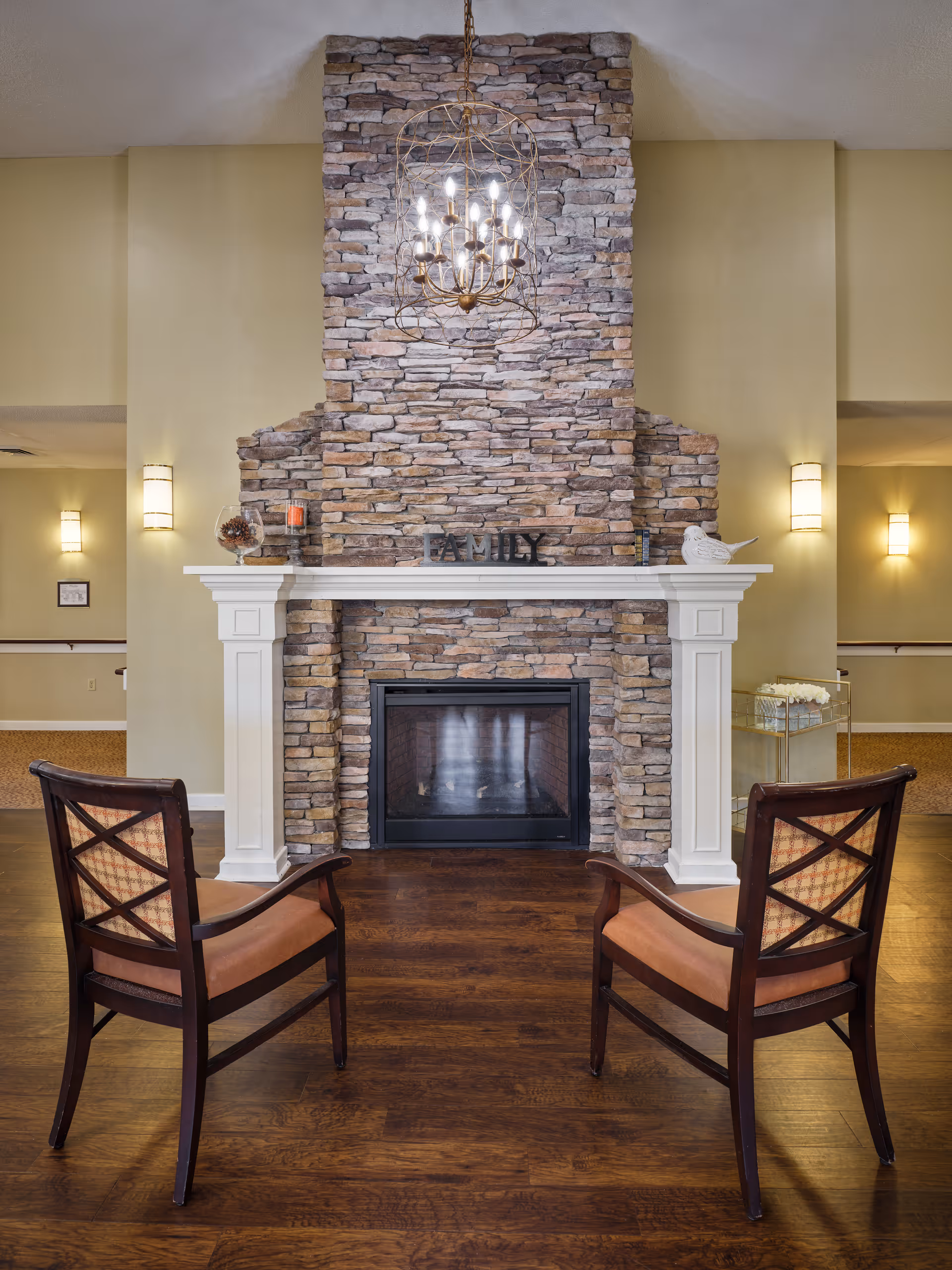 A cozy interior space featuring a stone fireplace with a white mantel. On the mantel, there are decorative items including a glass bowl with pinecones, a candle, a small sign that reads 'FAMILY', and a white bird figurine. Two wooden chairs with cushioned seats face the fireplace on a polished wooden floor. The walls are painted a soft beige color and there are wall sconces providing warm lighting. A chandelier hangs above the fireplace.