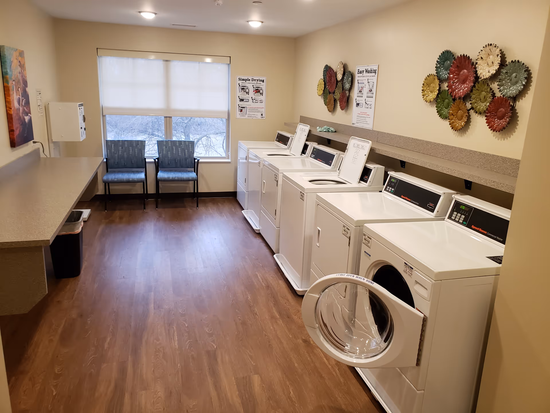 A clean laundry room with a row of washers and dryers, a folding counter, two chairs by a window, and decorative wall art.