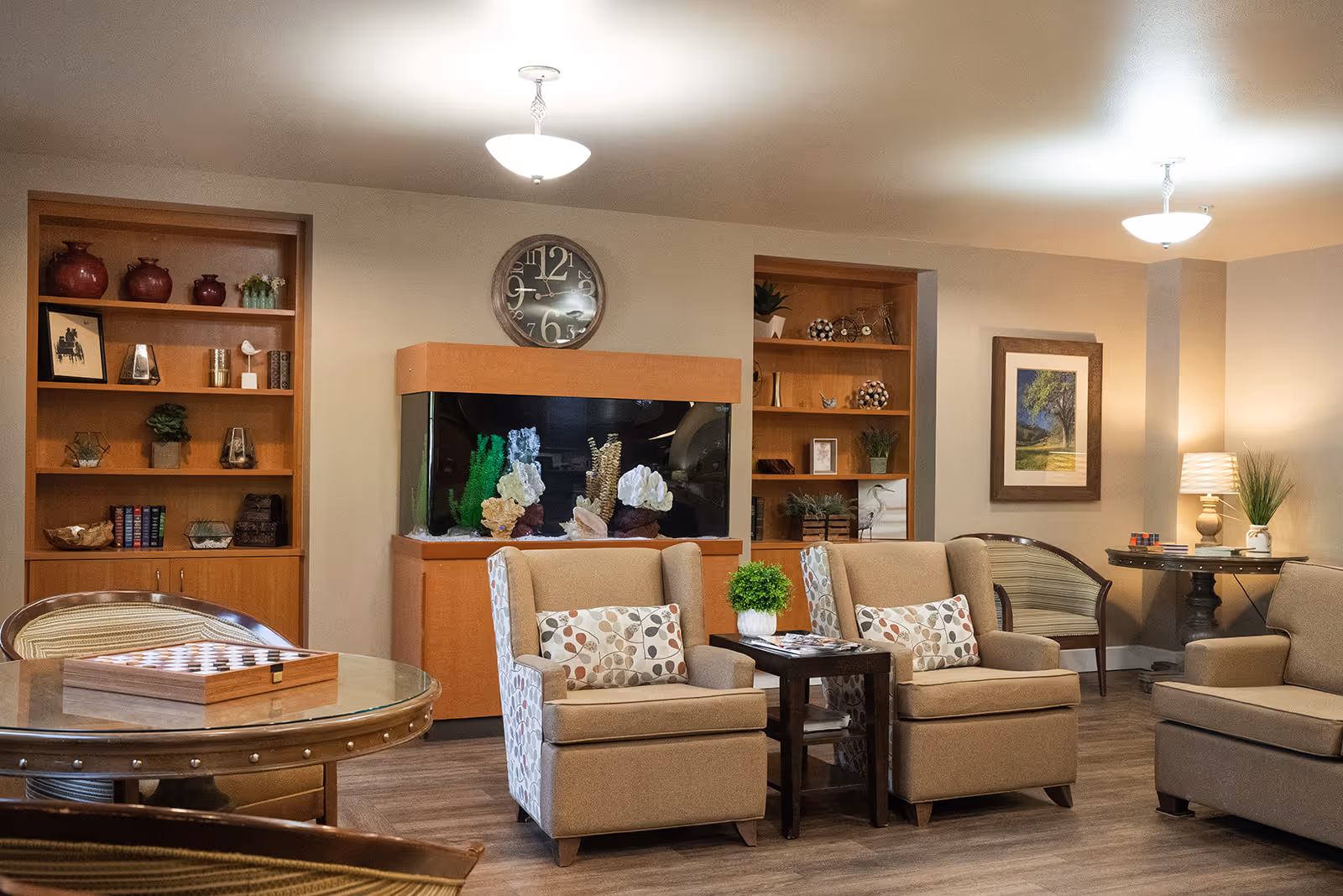 A cozy living room area with beige armchairs featuring patterned cushions, a small wooden side table with a green plant, a large fish tank built into the wall, wooden bookshelves with decorative items, a round table with a checkerboard game, and warm lighting from ceiling fixtures and a table lamp.
