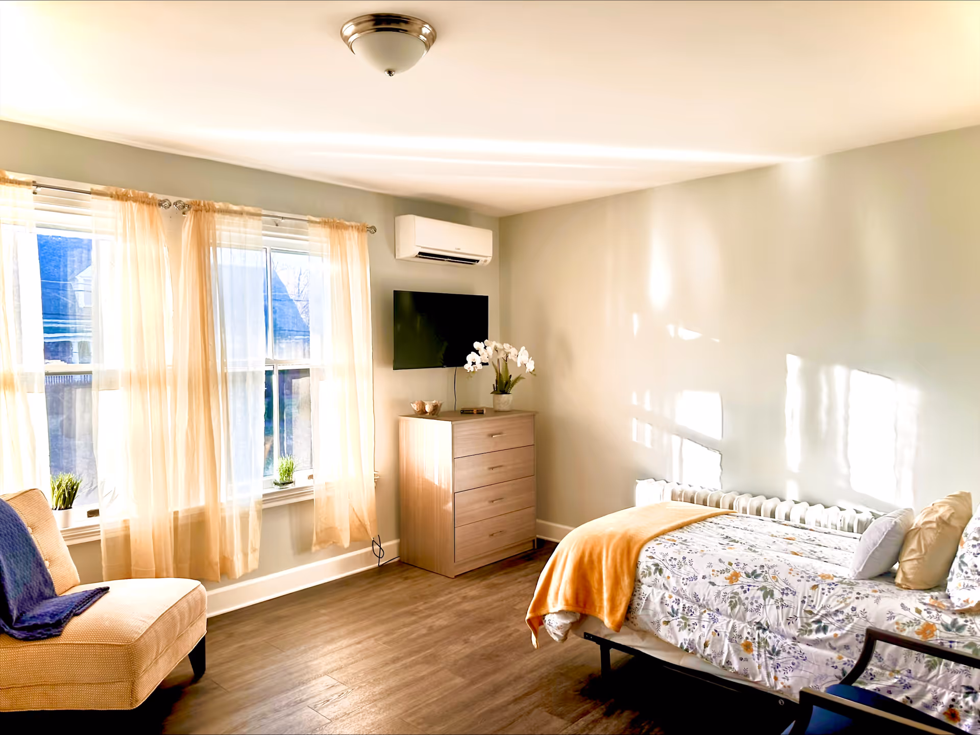 A bright and cozy bedroom in a senior living community featuring a single bed with floral bedding and an orange throw blanket, a wooden dresser with a potted orchid and a mounted flat-screen TV above it, a beige armchair with a blue throw blanket, and large windows with sheer curtains allowing natural light to fill the room.
