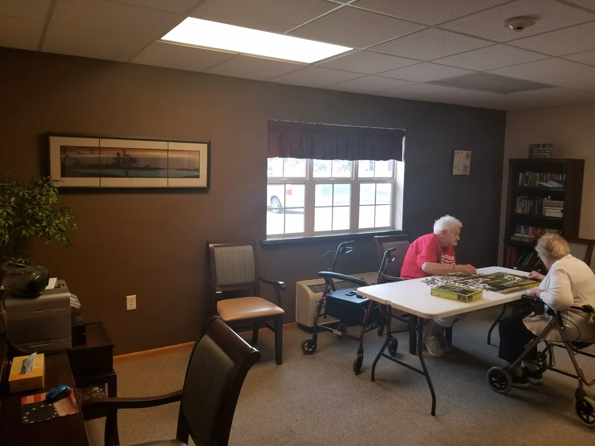 Two elderly women sit at a table working on a puzzle in a senior living community activity room.