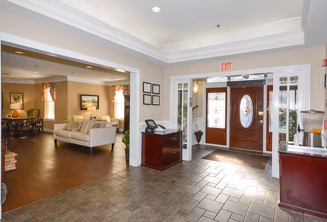 Interior view of a senior living facility entrance area with a tiled floor, a wooden front door with decorative glass panels, and an exit sign above. To the left is a living room area with a sofa, chairs, framed pictures on the walls, and windows with curtains allowing natural light. There is a small wooden cabinet with a phone on it near the entrance.