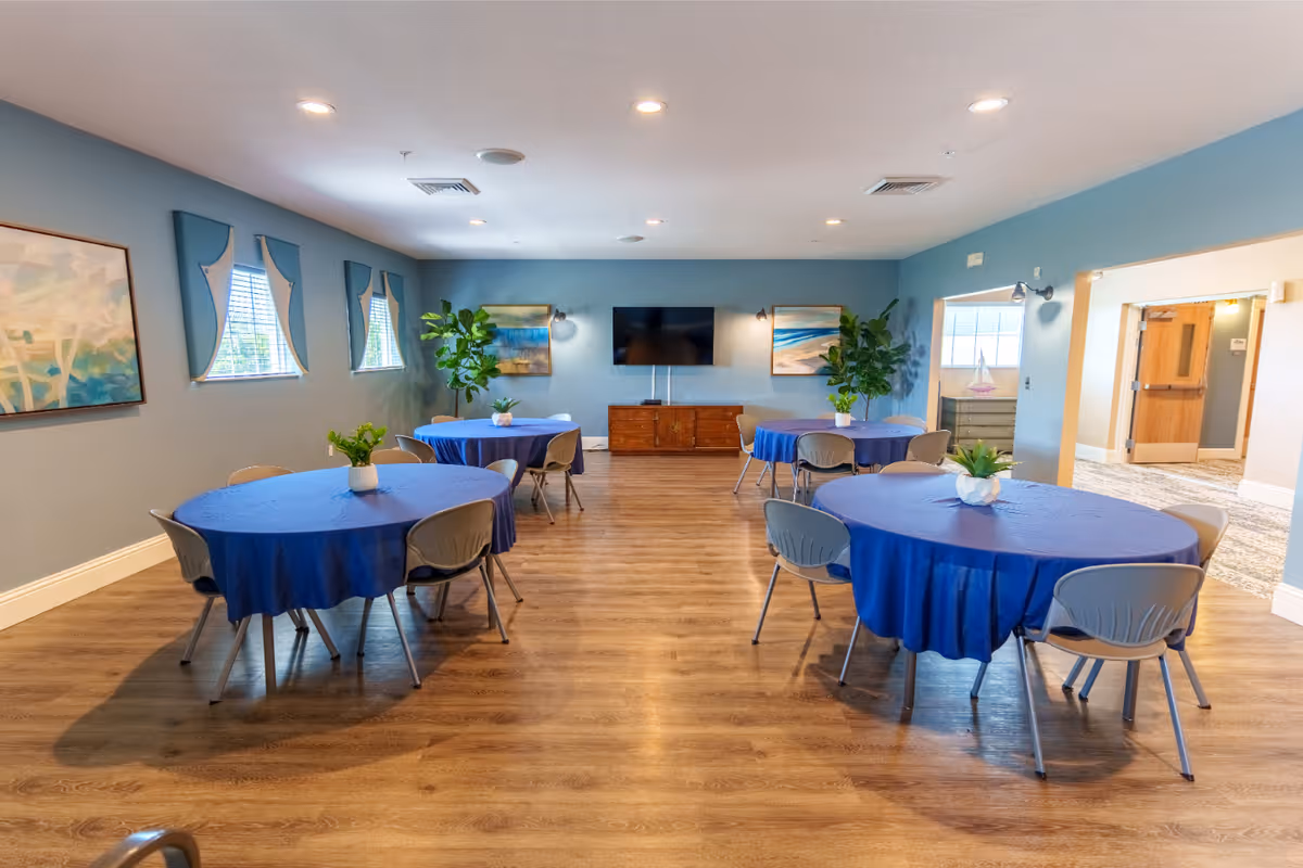 A spacious room with four round tables covered in blue tablecloths, each surrounded by gray chairs. The room has light blue walls with three windows on the left side, decorated with blue valances. There are two large green plants near the back wall, which also features a flat-screen TV mounted above a wooden cabinet and two framed paintings. The floor is wooden, and the room is well-lit with recessed ceiling lights.