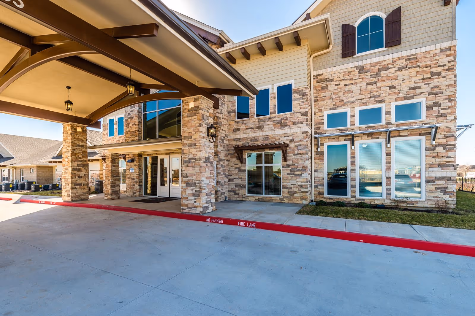 Exterior view of a senior living facility entrance with stone and beige siding, large windows, and a covered drop-off area supported by stone pillars. The driveway has a red curb marked 'No Parking Fire Lane'.