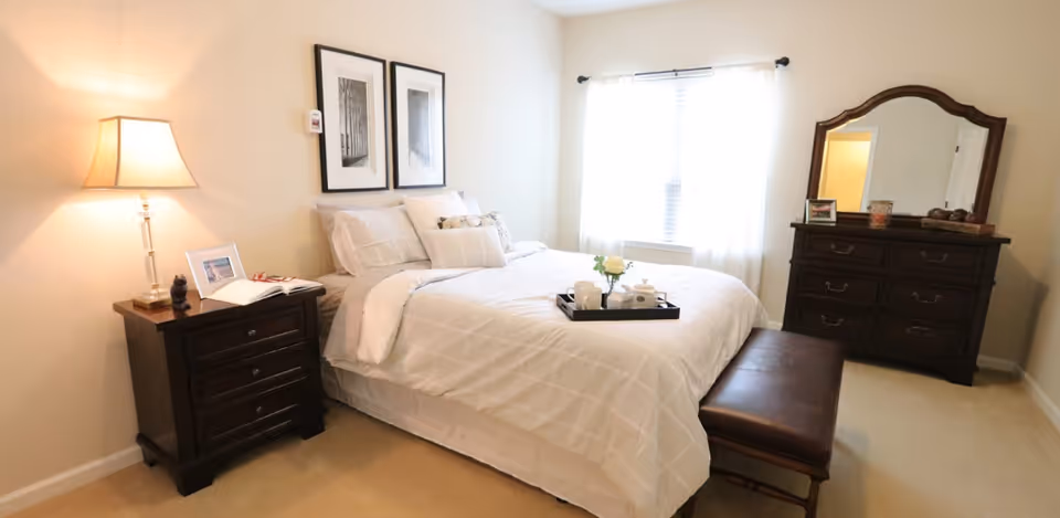 A well-lit bedroom with a neatly made bed featuring white bedding and multiple pillows. There is a dark wooden nightstand with a lamp, a framed photo, and an open book on the left side of the bed. Above the bed are two framed black and white pictures. On the right side of the room, there is a dark wooden dresser with a large mirror and decorative items on top. A brown bench is positioned at the foot of the bed. The room has beige walls and carpet, and a window with white curtains allowing natural light to enter.