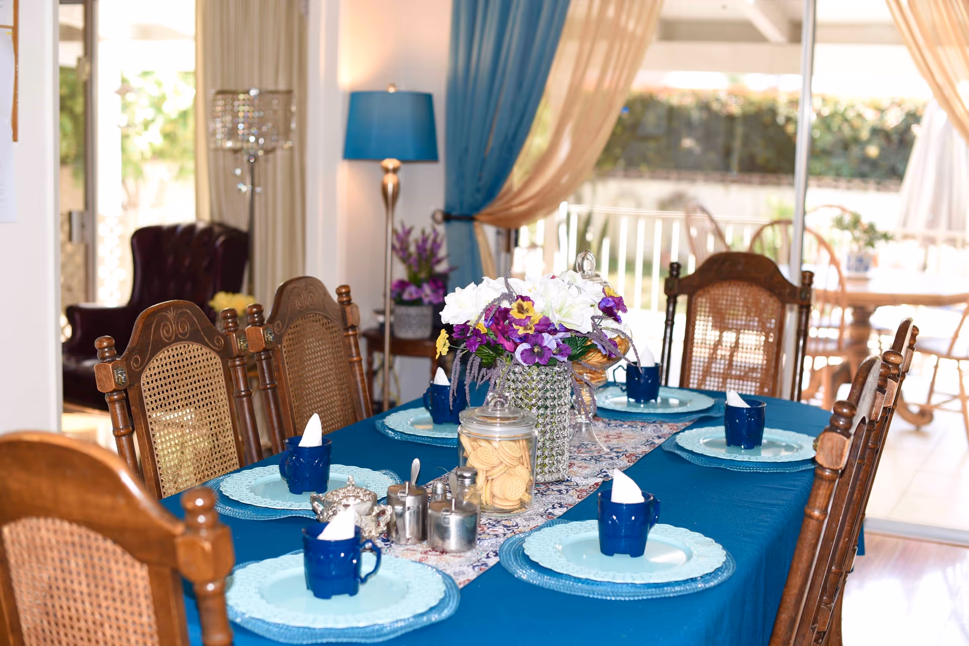 A dining table set for six with a blue tablecloth, floral centerpiece, and place settings in a bright room with sliding glass doors.