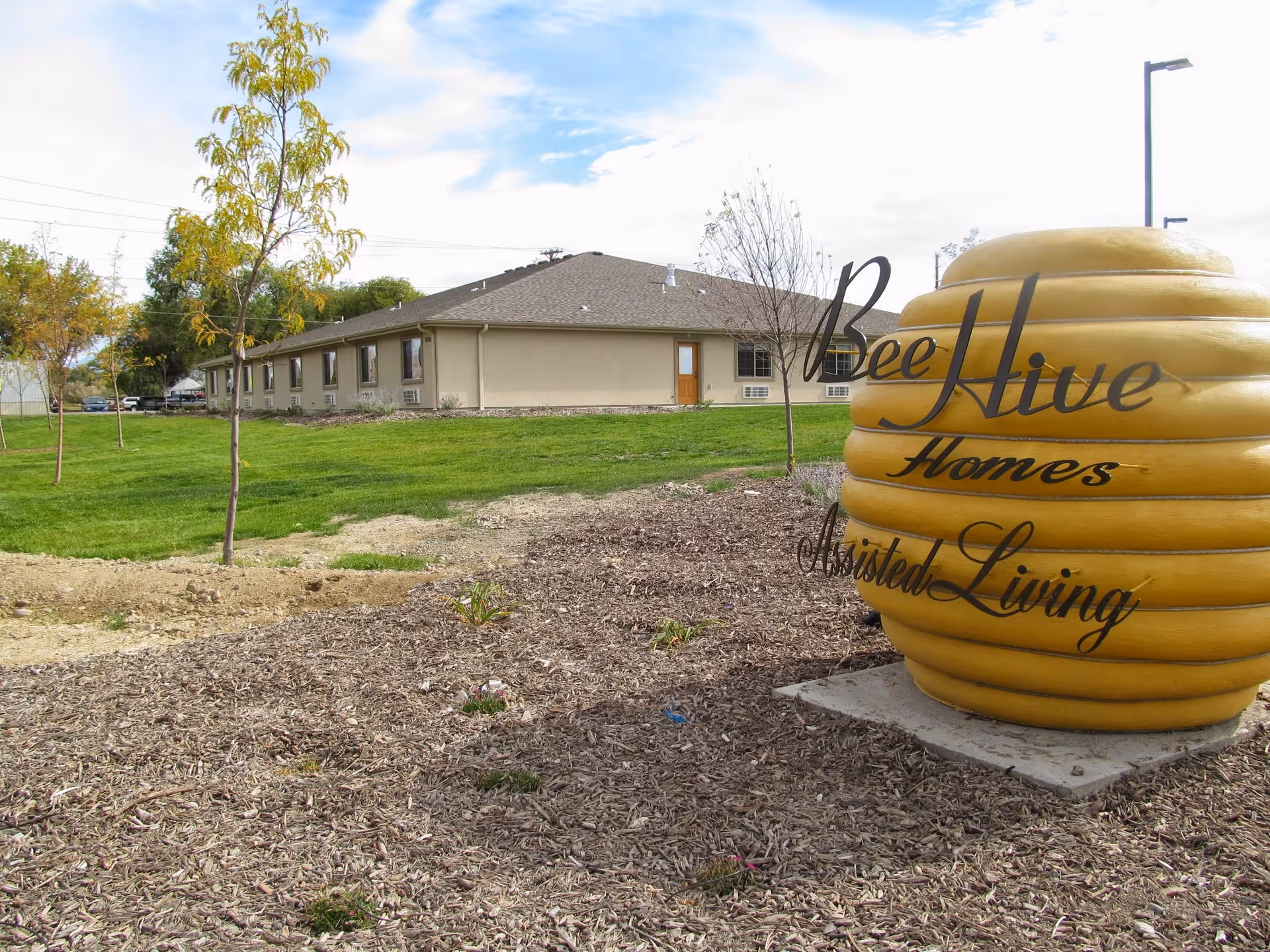 Outdoor view of BeeHive Homes Assisted Living facility showing a single-story beige building with a brown roof, surrounded by green grass and young trees. In the foreground, there is a large yellow beehive-shaped sculpture with the text 'BeeHive Homes Assisted Living' written on it.