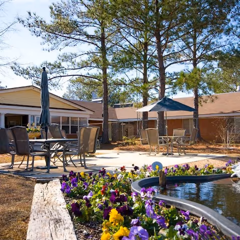 Outdoor patio area at Four County Health & Rehabilitation with tables, chairs, umbrellas, tall pine trees, and a flower bed with colorful pansies next to a small pond.