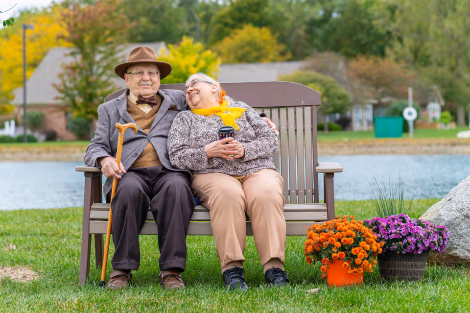 An elderly couple sitting closely together on a wooden bench by a lake, with the man holding a cane and the woman holding a cup. They are outdoors with green grass, colorful flowers in pots, and trees with autumn foliage in the background.