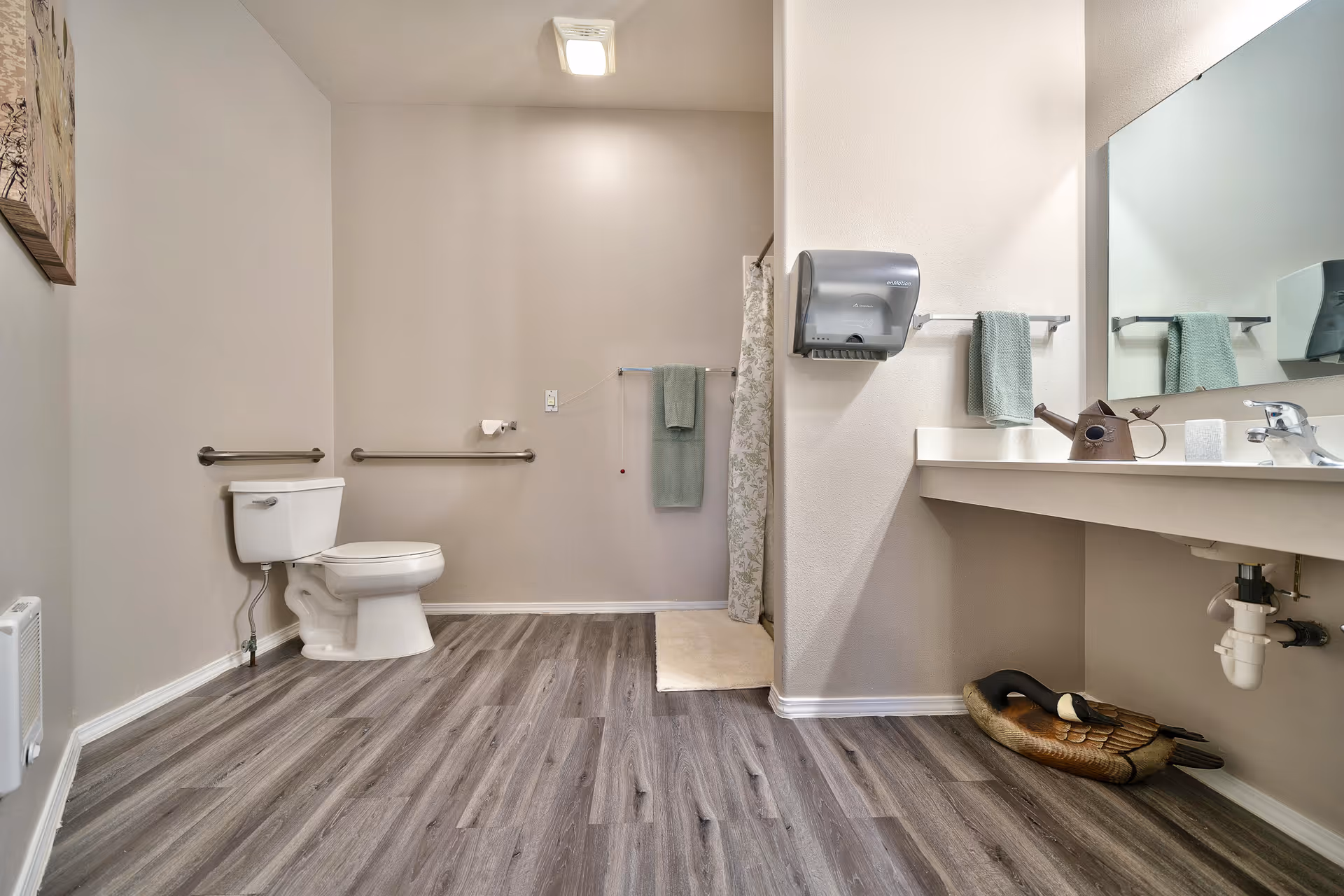 A spacious senior living bathroom with a white toilet, grab bars on the walls, a towel rack with green towels, a shower area with a floral curtain, a large mirror above a countertop sink, a wall-mounted paper towel dispenser, and a decorative wooden duck on the floor under the sink. The floor has wood-like vinyl planks and the walls are painted light beige.