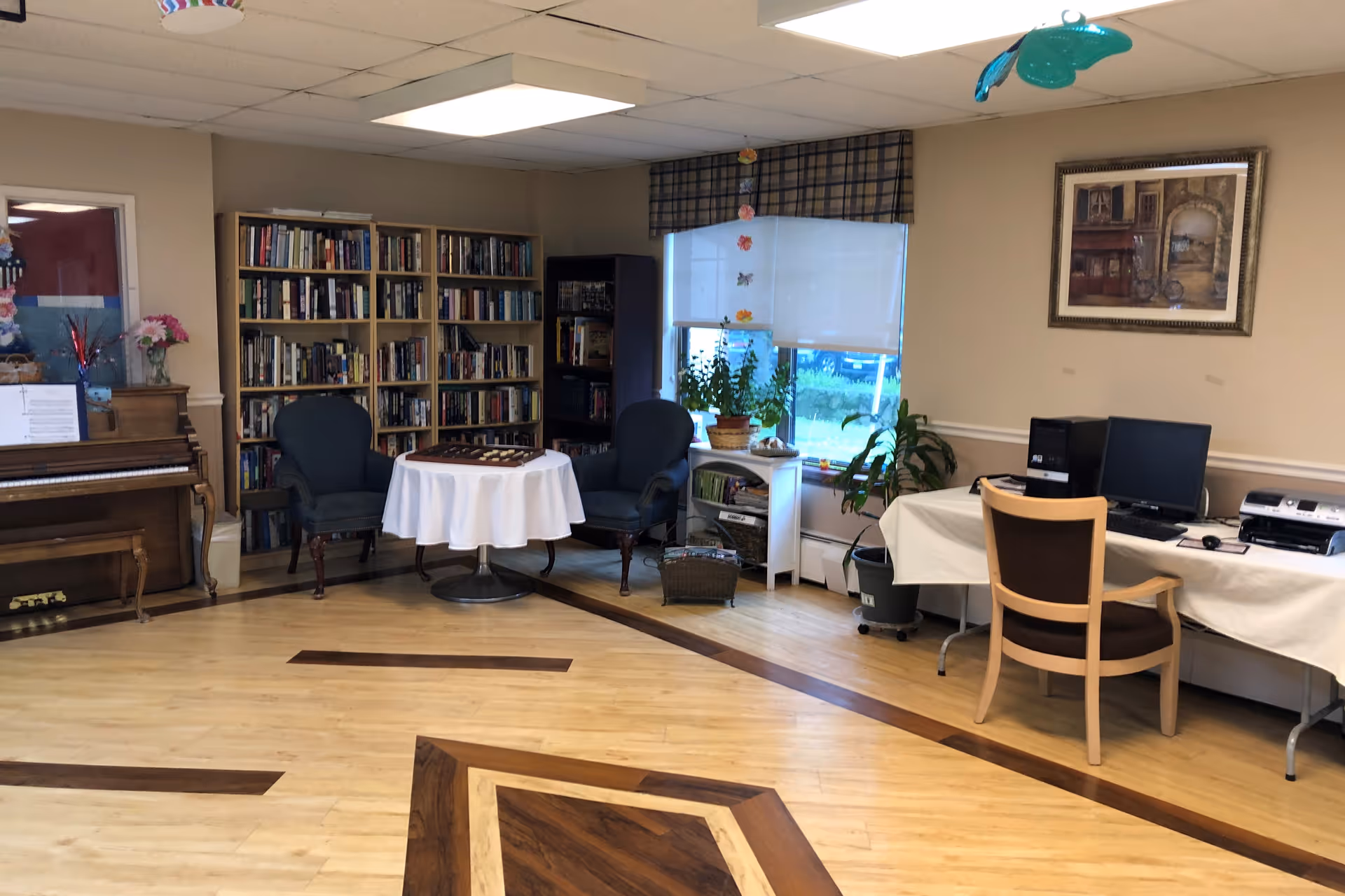 A cozy room with wooden flooring featuring a piano on the left, a bookshelf filled with books, two armchairs around a small round table with a white tablecloth, a window with plants on a small white shelf, and a desk with a computer and printer on the right. The walls are beige with a framed picture hanging above the desk.