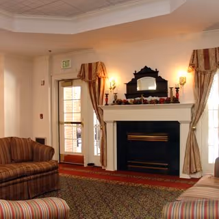 Cozy living room area with a fireplace decorated with candles and garland. There are two striped upholstered sofas facing each other, a patterned carpet, and large windows with curtains letting in natural light.