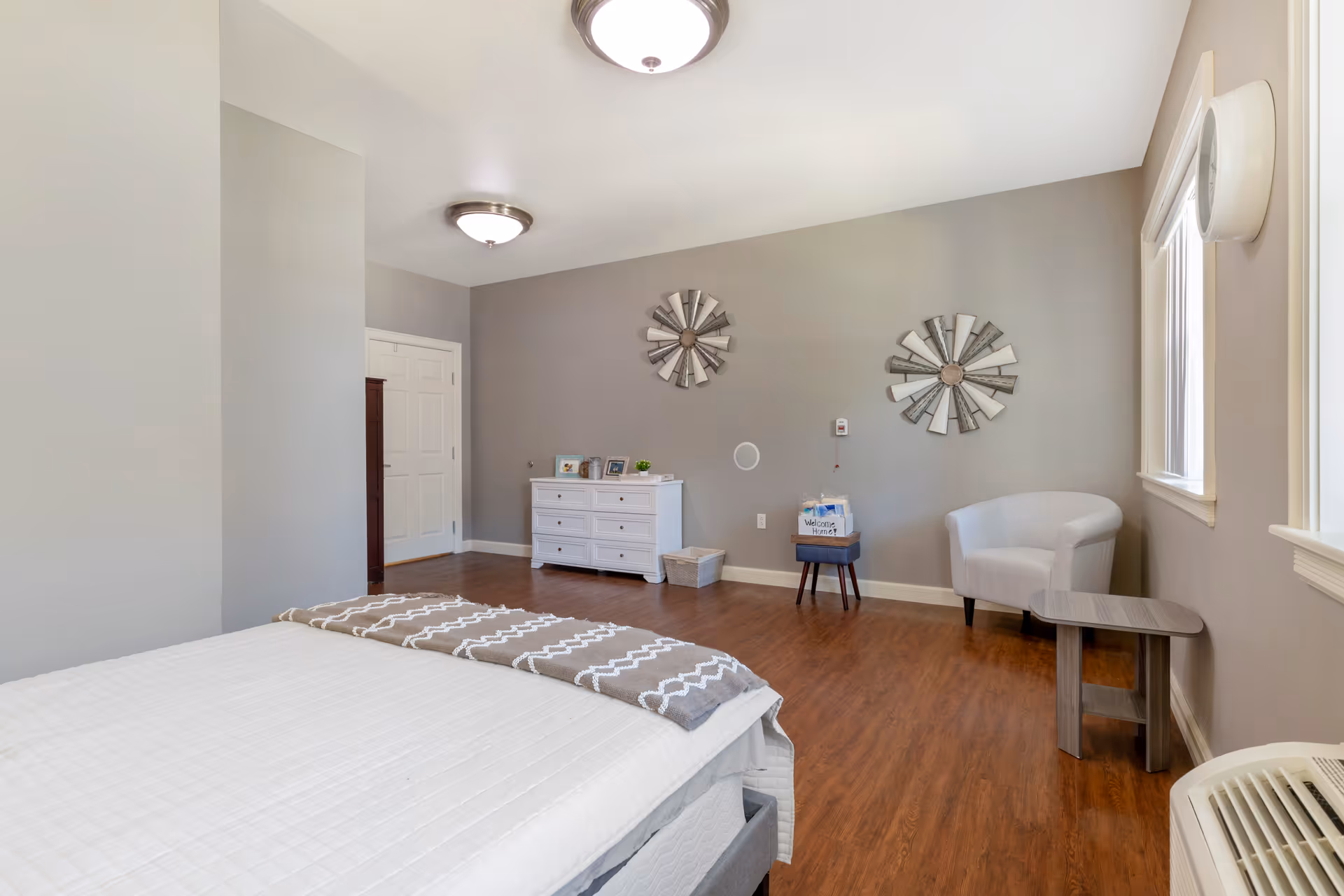 A clean and bright bedroom with a bed covered in a white quilt and a patterned throw blanket. The room has wooden flooring, light gray walls, and two decorative wall hangings above a white dresser. There is a white armchair and a small wooden side table near two windows letting in natural light. Ceiling lights illuminate the room.