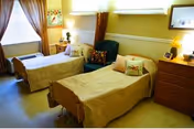 A well-lit room with two single beds covered in beige bedspreads, each with a decorative pillow. Between the beds is a green armchair with a floral cushion. The room has a window with curtains, a wooden dresser with a lamp and framed pictures, and light-colored walls with a chair rail.