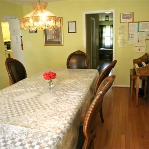 A dining room with a rectangular table covered with a patterned tablecloth and a small vase with red flowers in the center. Four wooden chairs with dark cushioned seats surround the table. A chandelier with multiple lights hangs above the table. The walls are painted light yellow, and there is a doorway leading to another room with a window and curtains. Various framed pictures and notices are on the walls, and a wooden piano is visible on the right side.