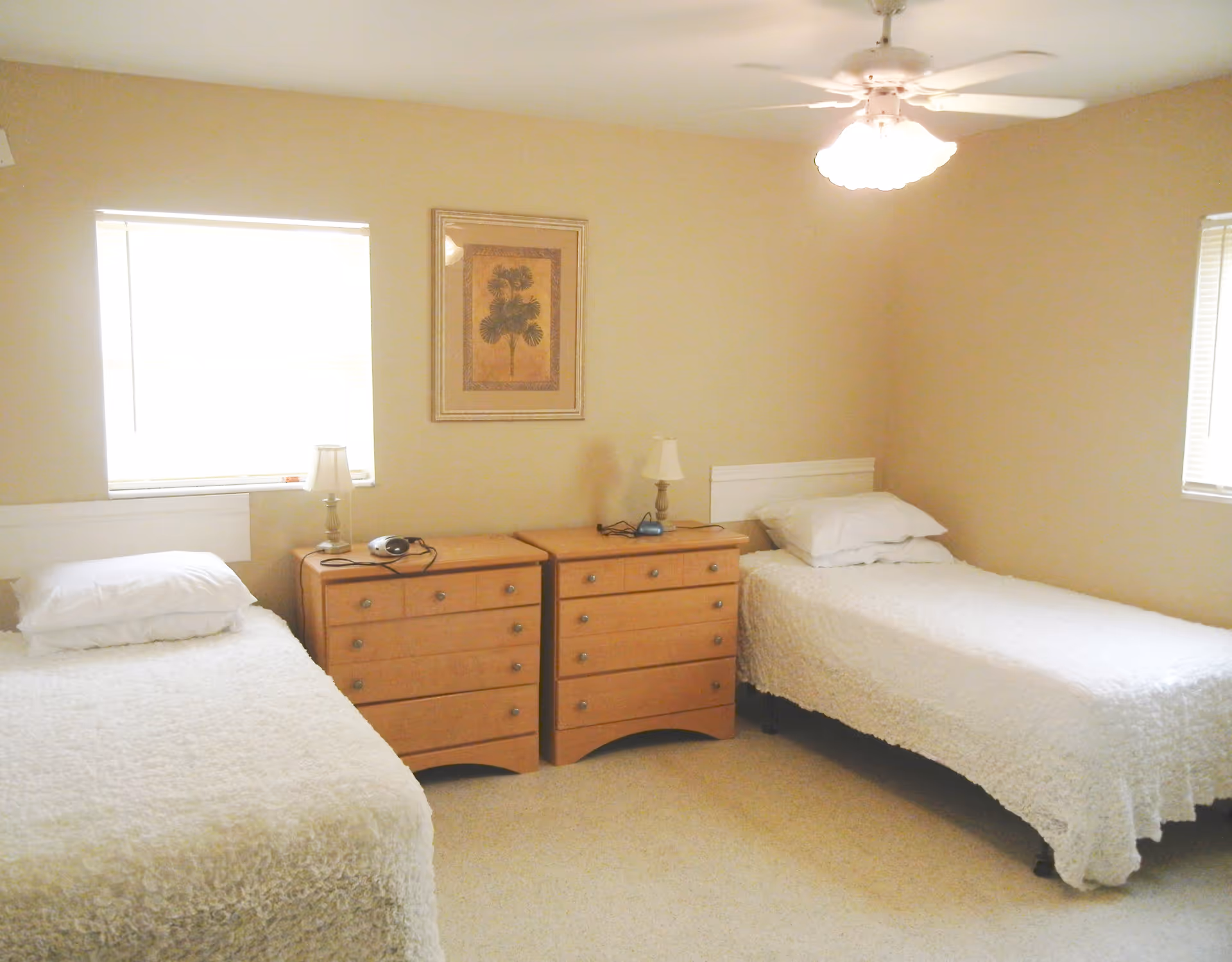 A bedroom with two single beds covered in white textured bedspreads, each with a pillow. Between the beds are two wooden dressers with lamps and small electronic devices on top. The walls are beige, with two windows covered by blinds and a framed picture hanging above the dressers. A ceiling fan with a light fixture is visible above.
