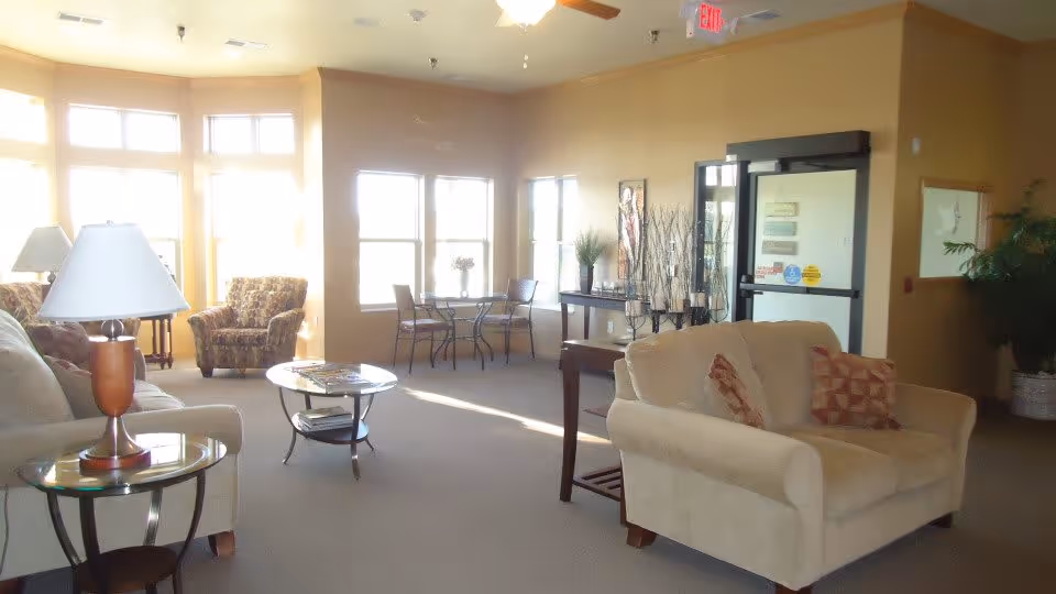 A bright and spacious living room area in an assisted living facility with beige walls and carpet. The room features a beige loveseat with patterned cushions, a glass-top side table with a lamp, a round coffee table, two armchairs near large windows, and a small dining table with chairs by the windows. There is a console table with decorative items and plants near a glass door with exit signs above.