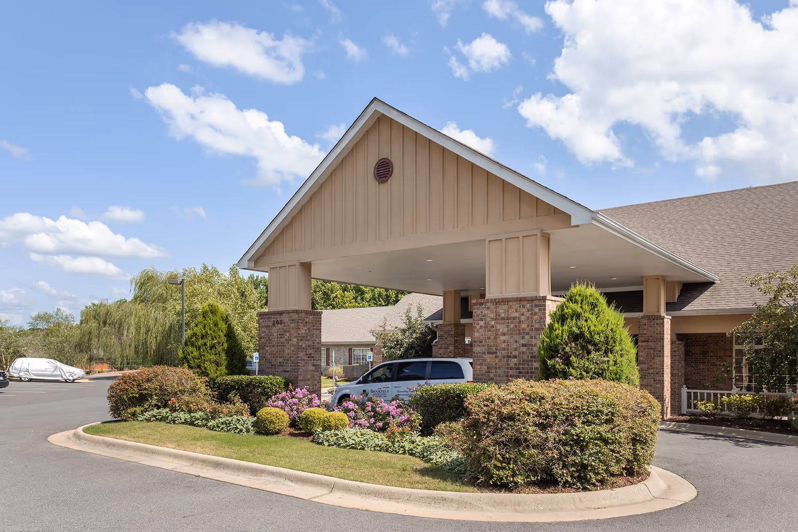 Entrance of Brookfield Assisted Living and Memory Care facility with a covered drop-off area supported by brick pillars, surrounded by well-maintained landscaping including bushes and flowering plants, under a partly cloudy blue sky.