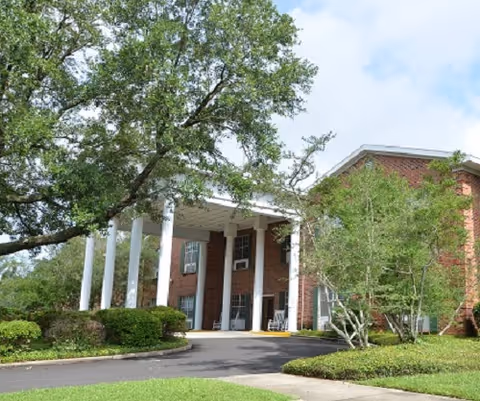 Brick senior living facility with white columns, a covered entrance, and surrounding trees.
