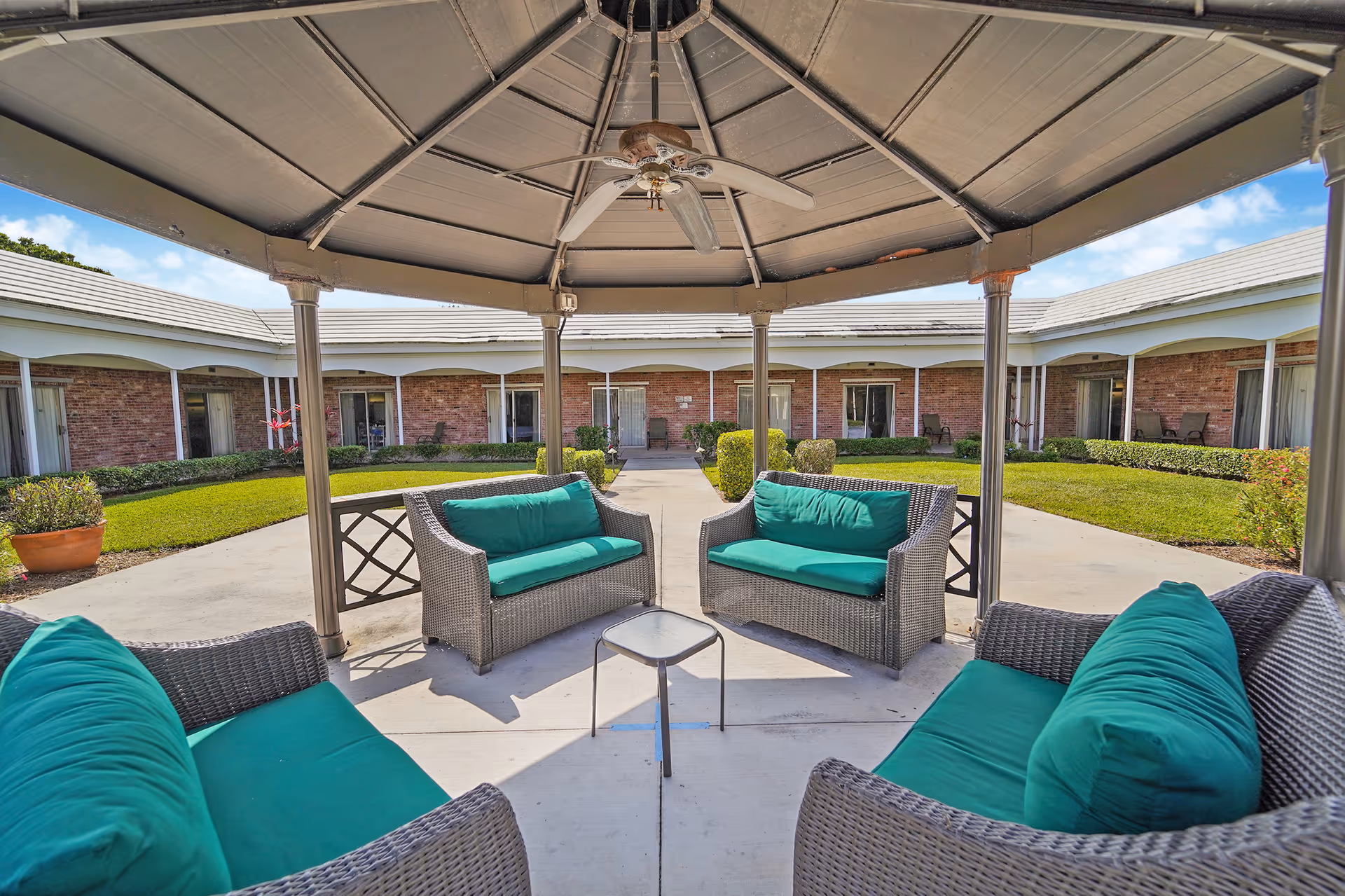 Outdoor seating area under a gazebo with four wicker sofas featuring teal cushions arranged around a small glass table, surrounded by a courtyard with green grass, shrubs, and a brick building with multiple doors and windows.